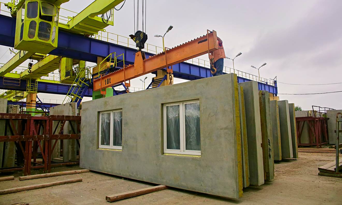 A crane lifts concrete modular wall panels with windows at a construction site.